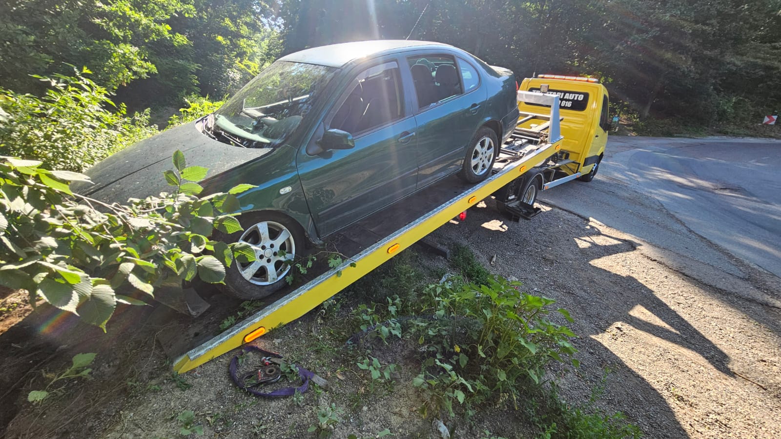 Autoturism verde cu roata rupta scos din padure cu platforma galbena Traian TKD, echipata cu sistem hidraulic.