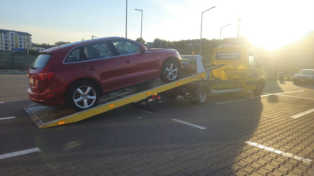 Serviciul nostru de **tractari auto Iasi pret** avantajos în acțiune: platforma galbenă Traian TKD încarcă în siguranță un SUV Audi Q5 roșu într-o parcare, la apus.
